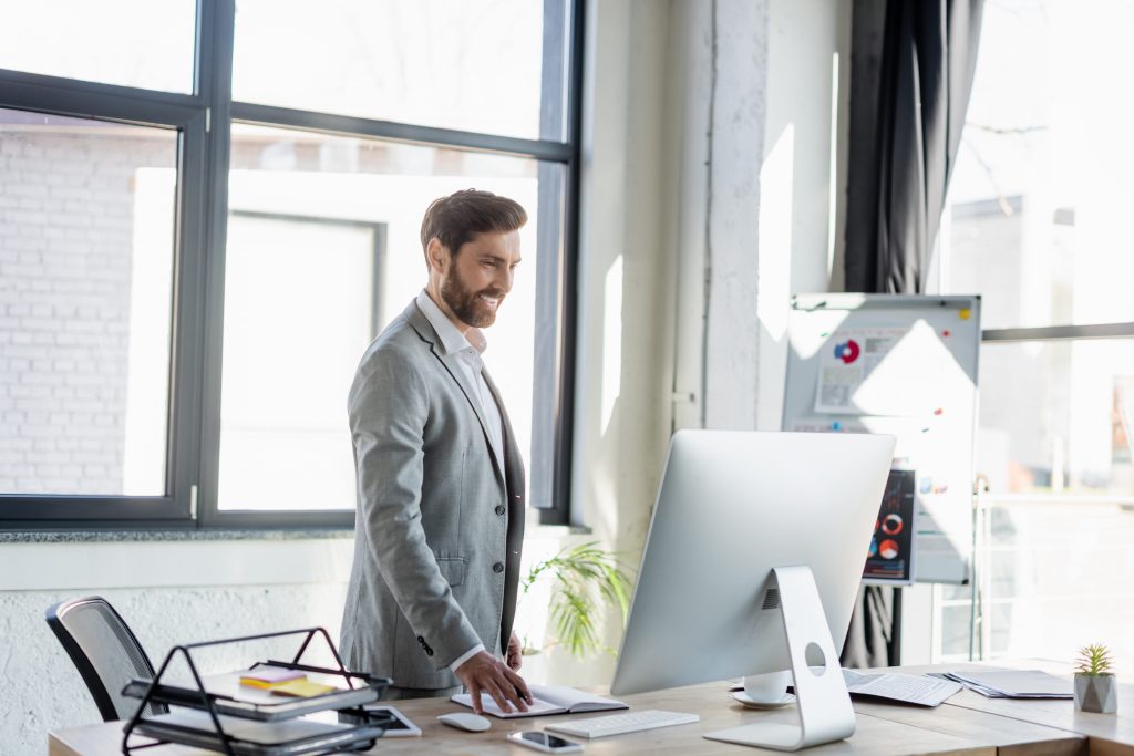 Business professional standing at a desk in a bright modern office, reviewing information on a large computer monitor while managing operations in a productive and technology-driven work environment.
