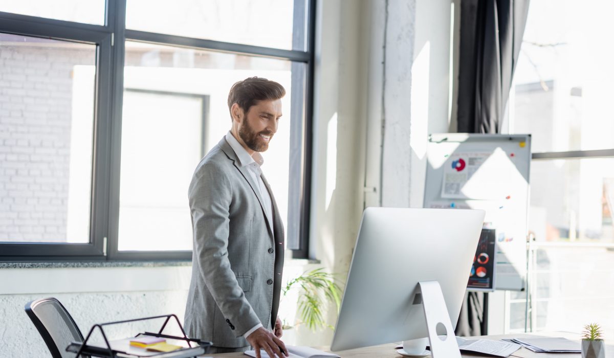 Business professional standing at a desk in a bright modern office, reviewing information on a large computer monitor while managing operations in a productive and technology-driven work environment.